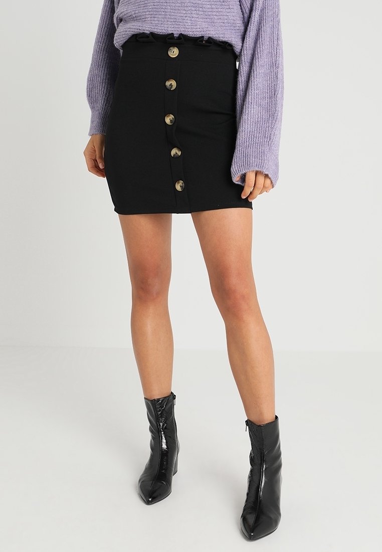 Femme portant une mini-jupe noire à boutons sur le devant, un pull lavande à manches longues et des bottines noires brillantes, debout devant un fond uni.