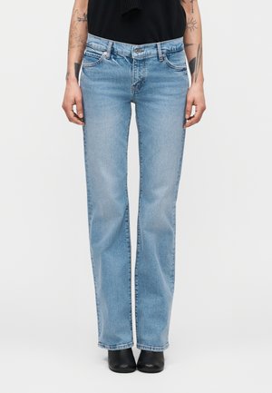 Personne portant un jean bleu clair à jambes droites et des bottines noires, visible de la taille aux pieds sur un fond uni.