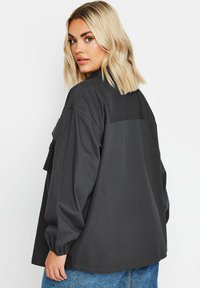 Chemise surdimensionnée grise foncée avec manches longues, poche poitrine boutonnée et tissu lisse légèrement texturé. La vue de dos montre une coupe décontractée.