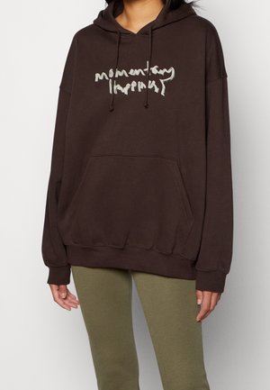 Personne portant un sweat à capuche ample marron foncé avec poche avant et texte beige illisible, associé à des leggings verts olive.