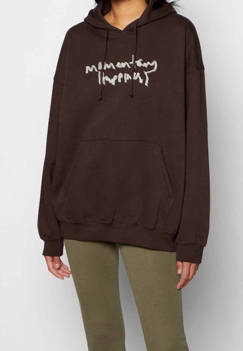 Personne portant un sweat à capuche ample marron foncé avec poche avant et texte beige illisible, associé à des leggings verts olive.