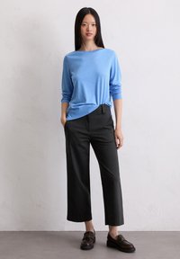 Frau mit langen, glatten Haaren, die ein hellblaues Langarmoberteil, schwarze, cropped, weit geschnittene Hosen und dunkelbraune Slipper trägt und vor einer weißen Wand steht.