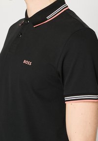 Polo noir avec col et bordures de manches rayés rose, blanc et noir, avec le logo "BOSS" rose sur la poitrine.
