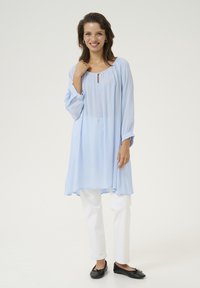 Tunique bleu clair à manches longues, décolleté en forme de clé et silhouette fluide, assortie à un pantalon blanc et des ballerines noires.
