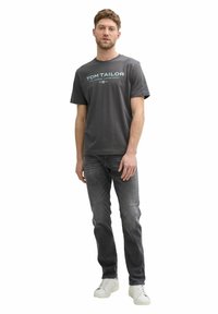 Gray cotton t-shirt featuring "TOM TAILOR" logo in teal, paired with dark jeans and white sneakers. Model stands in a neutral pose.
