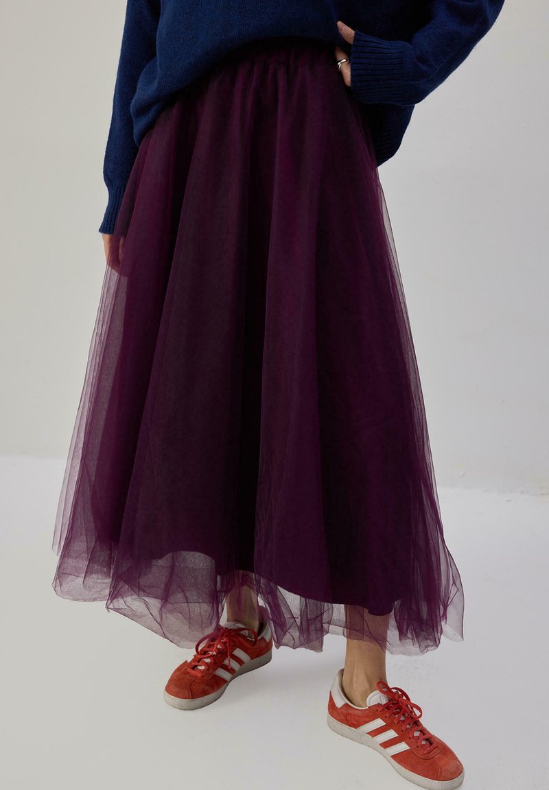 Gonna di tulle viola con vita arricciata, texture a strati, abbinata a sneakers arancioni e bianche. Maglione blu scuro.