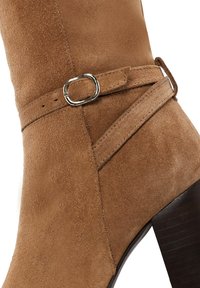 Stivale alla caviglia in suede marrone con dettaglio a fibbia, superficie texturizzata e tacco alto affusolato. Il design include un accento con cinturino diagonale.