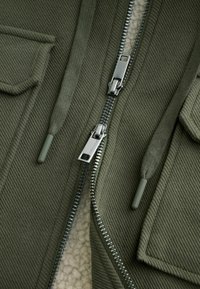 Alyvinės žalios spalvos tekstūruota striukė su dviem užtrauktukais, reguliuojamu gobtuvu ir matoma minkšta balta vilna viduje.