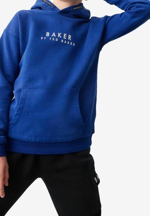 Dziecko ubrane w niebieską bluzę z kapturem z napisem „BAKER BY TED BAKER” i czarne spodnie z boczną kieszenią na białym tle.