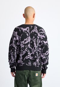 Pulover tricotat negru cu un model abstract violet, având gât rotund și mâneci cu elastic. Purtați cu pantaloni din catifea cord cu un logo.