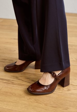 Escarpins en cuir verni marron avec un bout carré et une bride de cheville ornée d'une boucle circulaire. Le talon est épais et texturé.