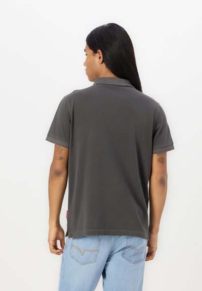 Polo shirt en coton gris avec col et manches courtes. Présente une texture subtile et une étiquette de marque en bas. Porté avec un jeans bleu clair.