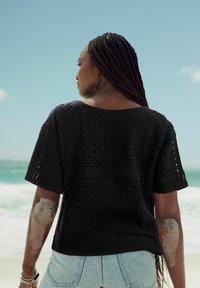 Sorte strikket top med korte ærmer, der har et struktureret mønster, parret med højtaljede denimshorts, sand på armen, havbaggrund.