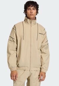 Μπεζ ζακέτα με φερμουάρ και ψηλό γιακά, μαύρες διαγραμμίσεις στους ώμους και ελαστικά μανίκια. Φέρει το λογότυπο της Adidas στο στήθος.