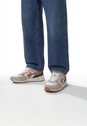 Personne portant un jean bleu et des baskets décontractées blanches, beiges et marron debout sur un sol blanc.