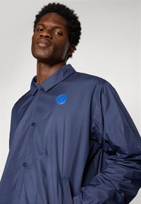 Young Black man wearing a navy blue button-up jacket with a round blue emblem on the chest, standing against a plain light background.