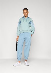 Lys blå hettegenser med grønne accenter og oversized ermer, kombinert med matchende joggebukser og hvite sneakers. Modell holder en svart vannflaske.
