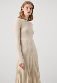 Beige bordás kötött ruha hosszú ujjal, formázott testen és plisszírozott szoknyával. Különlegessége a hamis nyak és a diszkrét fényesség.