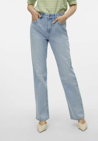 Jeans de tiro alto azul claro con piernas rectas y bolsillos frontales, combinados con una blusa texturizada verde y tacones beige de punta.