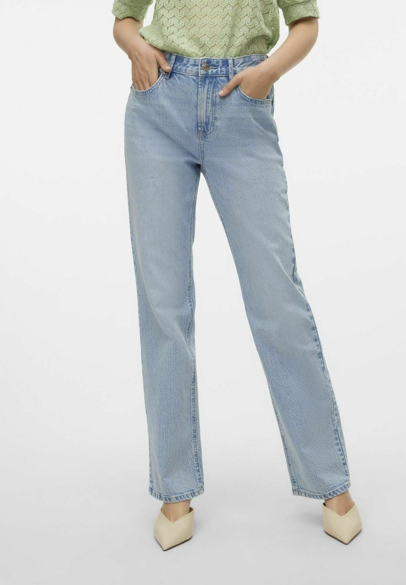 Jeans de tiro alto azul claro con piernas rectas y bolsillos frontales, combinados con una blusa texturizada verde y tacones beige de punta.