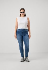 Λευκό αμάνικο τοπ, μπλε skinny τζιν, γκρι αθλητικά παπούτσια. Μοντέλο που φορά μαύρα γυαλιά ηλίου, standing against a gray background.