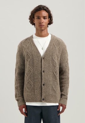 Man in een bruin gestructureerd knoopvest over een wit shirt en donkere broek, staande tegen een effen lichtgrijze achtergrond.