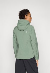 Lys grønn vanntett jakke med hette, glidelås og subtil merking. Glatt tekstur med en tettsittende design og lange ermer.