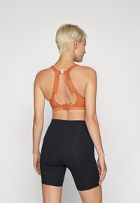Rustfarget sports-bh med racerback-design, justerbare stropper og krokfeste. Kombinert med sorte tettsittende shorts, glatt tekstur.