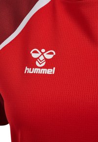 Rotes Sporttrikot mit weißem Hummel-Logo, das eine stilisierte Biene und Chevron-Muster auf einem strukturierten, atmungsaktiven Stoff zeigt.