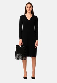 Robe noire à manches longues avec un décolleté en V, texture lisse. Le mannequin tient un sac à franges noir avec des clous argentés, associé à des talons noirs.