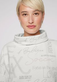 Sudadera de cuello cisne en gris claro con tejido texturizado. Presenta varios diseños en texto en gris oscuro y rosa suave. Ajuste casual y relajado.