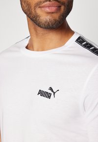 Valge lühikeste varrukatega T-särk, millel on must Puma logo ja brändinimi tikandiga rinnal; must aktsentkand koos õmbluste jaotustega.