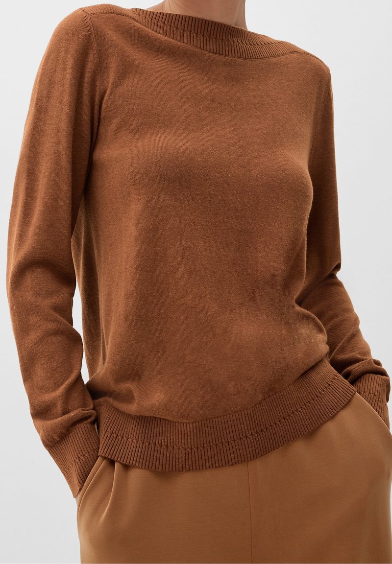 Personne portant un pull en maille brun à manches longues avec poignets et bas côtelés, associé à un pantalon marron assorti, mains dans les poches.