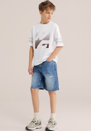 Dječak s kratkom kovrčavom kosom stoji noseći bijelu majicu s grafičkim motivom, traper kratke hlače, bijele čarape i sive tenisice, gledajući prema lijevo.