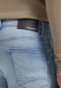 Ljust blå denimjeans med medelhög midja, som har en svart läderlapp märkt "PURE PATH" och synlig sömnad. Bakfickans design är standard.