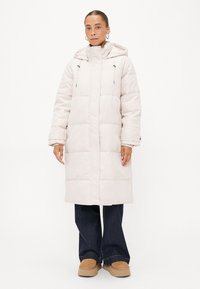 Lang, lichtgrijze gewatteerde jas met een hoge kraag, verstelbare trekkoordcapuchon en rechthoekige quilt. In combinatie met donkere denim en tan kleurige schoenen.