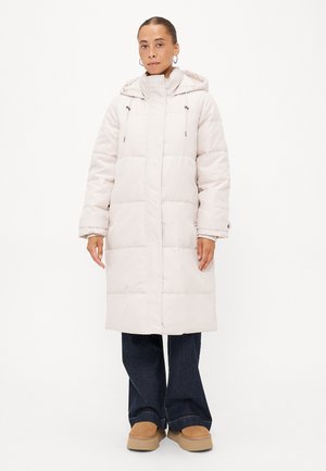 Manteau matelassé long et gris clair avec un col haut, une capuche ajustable à cordon et un quilting rectangulaire. Associé à un jean foncé et des chaussures beige.