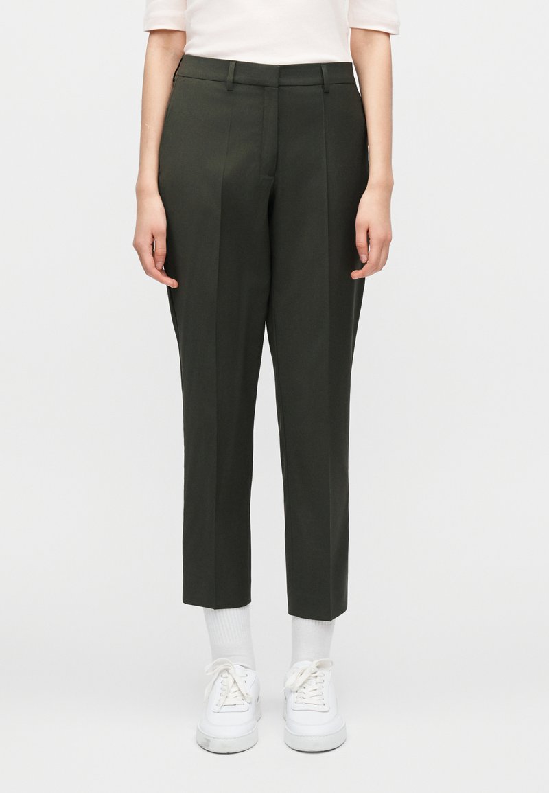 Άτομο που φοράει σκούρα πράσινα πρ οσαρμοσμένα παντελόνια μέχρι τον αστράγαλο, λευκές κάλτσες, λευκά αθλητικά παπούτσια και ανοιχτό ροζ μπλουζάκι, στέκεται μπροστά σε απλό φόντο.