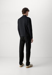 Zwarte zip-up jack met hoge kraag, zacht van textuur, gecombineerd met zwarte broek en beige schoenen, bekeken vanuit de achterkant.