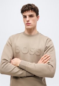 Νέος άνδρας με κοντά σγουρά μαλλιά, φοράει ένα μπεζ φούτερ με την επιγραφή "BOSS" στο εμπρόσθιο μέρος, με σταυρωμένα τα χέρια, σε απλό φόντο.