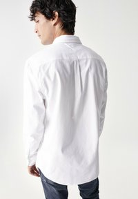 Camicia bianca in cotone con maniche lunghe, colletto a punta, cucitura della yoke posteriore e orlo arrotondato, indossata su jeans scuri.