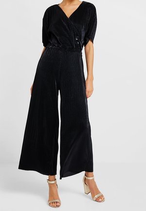 Woman wearing a black wide-leg jumpsuit with a V-neck, short sleeves, and beige high-heeled sandals standing on a white floor.