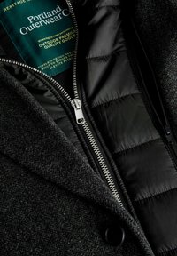 Grå yderjakke med tekstureret stof, med sort polstret inderside. Dobbelt lynlås med sølvfarvet metal og et grønt mærke synligt.