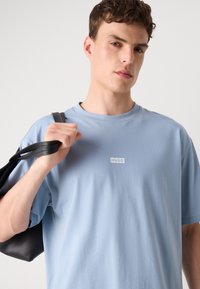 Ljusblå bomulls-t-shirt med rund hals och korta ärmar, prydd med en vit "HUGO"-logotyp på bröstet. Modellen håller en svart väska.