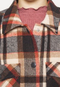 Nærbillede af en person iført en rød, brun, sort og cremefarvet plaid knapjakke over en ribbet, scallopkantet mørk pink top.