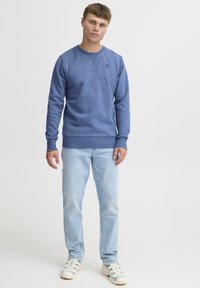 Blue sweatshirt with long sleeves, round neck, and ribbed hem; worn with light blue jeans and white sneakers. Simple logo detail on chest.