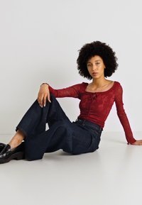 Κόκκινη λουλουδάτη μπλούζα από διχτυωτό υλικό με μακρύ μανίκι και κλείσιμο με κορδόνι, συνδυασμένη με φαρδιά τζιν παντελόνια από σκούρο τζιν. Λείο υλικό και χαλαρή εφαρμογή. Μαύρες μοκέτες.