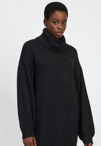 Schwarzes Strickkleid mit hohem Schalkragen, übergroßen Ärmeln und lockerer Passform. Hergestellt aus einem strukturierten Strickstoff.