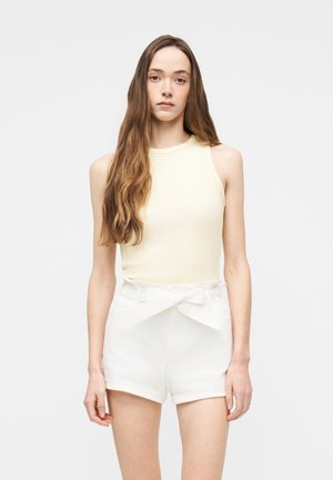 Jeune femme aux longs cheveux bruns portant un haut crème sans manches et un short blanc taille haute avec une ceinture nouée, debout devant un fond uni.