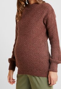 Těhotná žena v hnědém pleteném svetru s texturou a olivově zelených kalhotách, ukazující svůj těhotenský břicho.